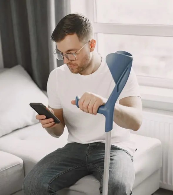 homme blessé sur son canapé avec un béquille, regardant son smartphone